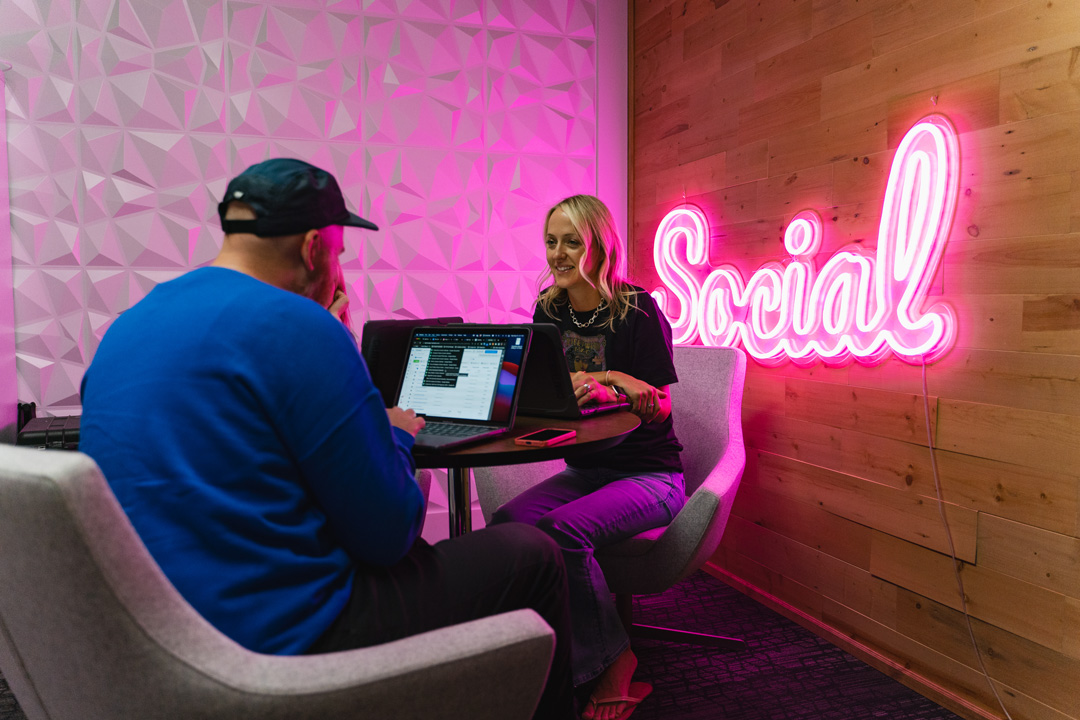 Mary sits talking to Austin, a neon sign is behind them reading Social on a wood panel wall
